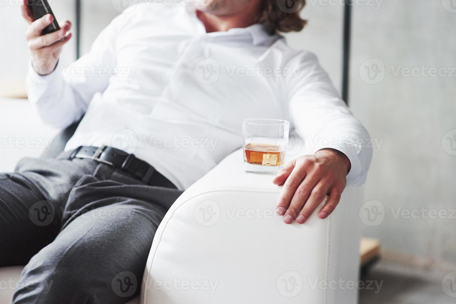 homme au bureau porter assis sur le canapé avec un téléphone à la main et du whisky sur le côté photo