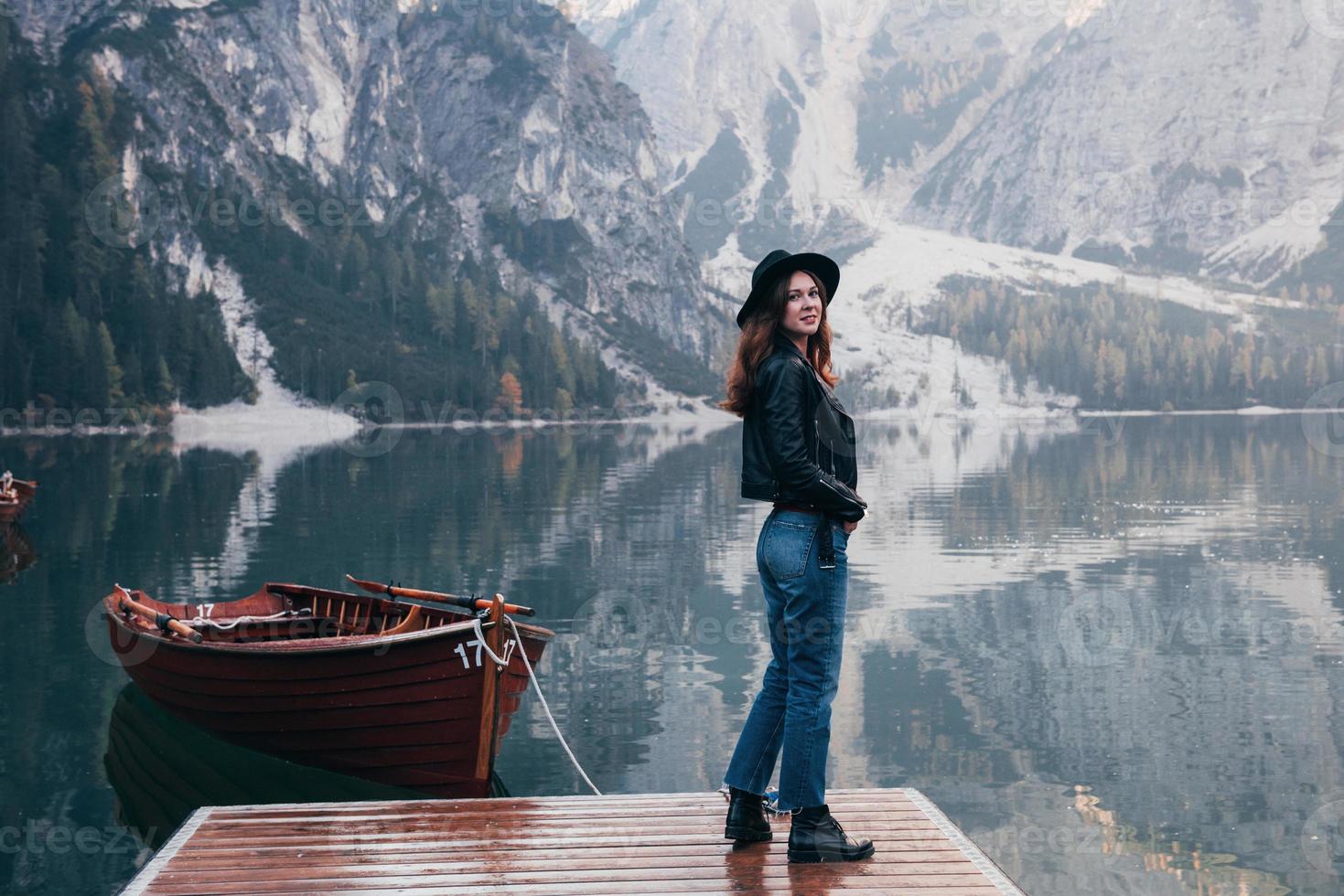 la fille est retournée. femme au chapeau noir profitant d'un paysage de montagne majestueux près du lac avec des bateaux photo