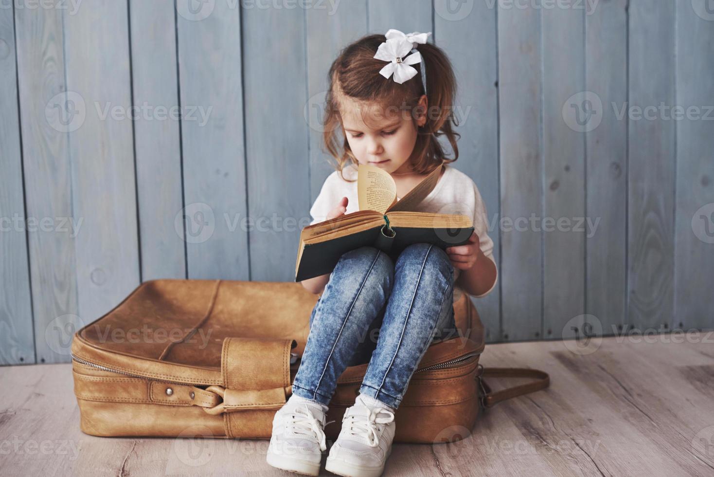 prêt à grand voyage. heureuse petite fille lisant un livre intéressant portant une grosse mallette. concept de liberté et d'imagination photo