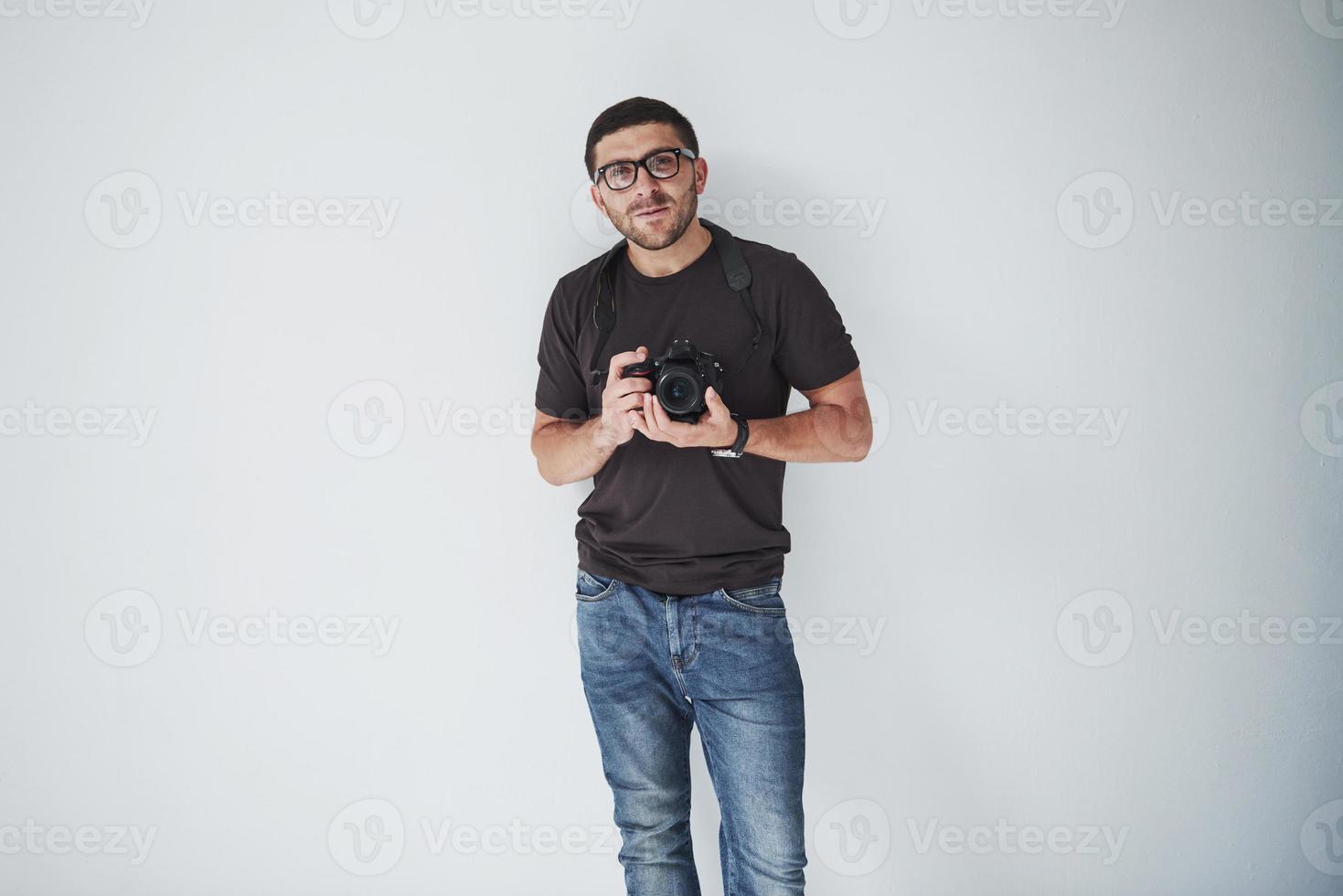 un jeune homme hipster dans les oculaires tient un appareil photo reflex numérique dans les mains debout sur un fond de mur blanc