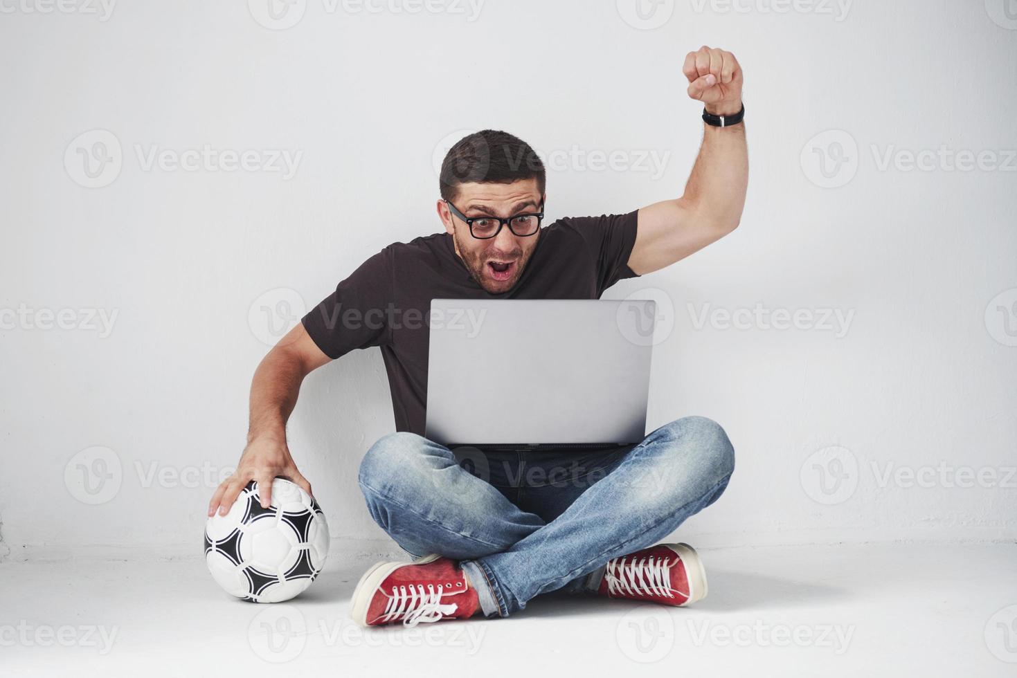 fan de football excité avec un ballon de football isolé sur fond blanc. il est assis par terre et regarde les résultats du match et les meilleurs moments du match sur l'ordinateur portable. photo