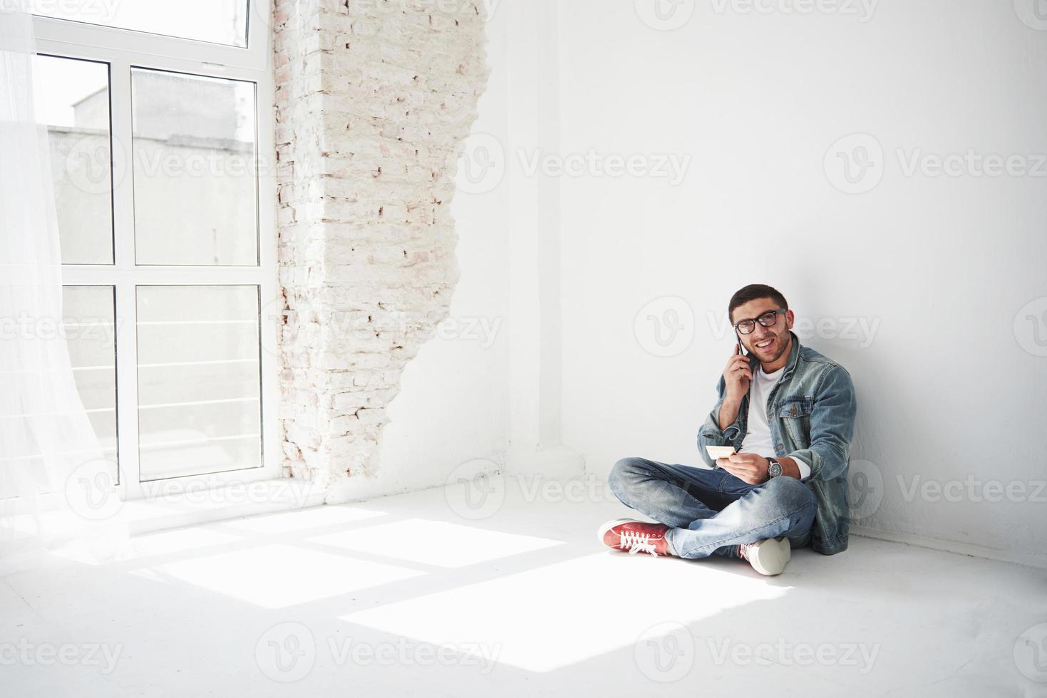 un gars en vêtements décontractés est assis à la maison dans un appartement vide, tenant une carte de crédit et appelant au téléphone. peut-être est-il un nouveau résident et n'a-t-il pas encore acheté de meubles. photo