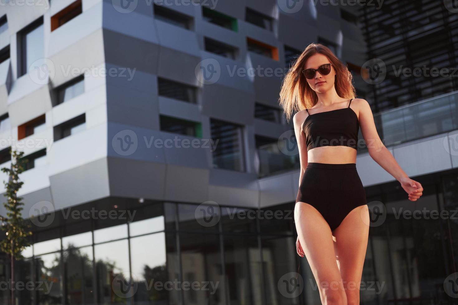 sexy jeune femme à lunettes de soleil et maillot de bain noir est debout sur le fond des bâtiments de la ville pendant le coucher du soleil photo