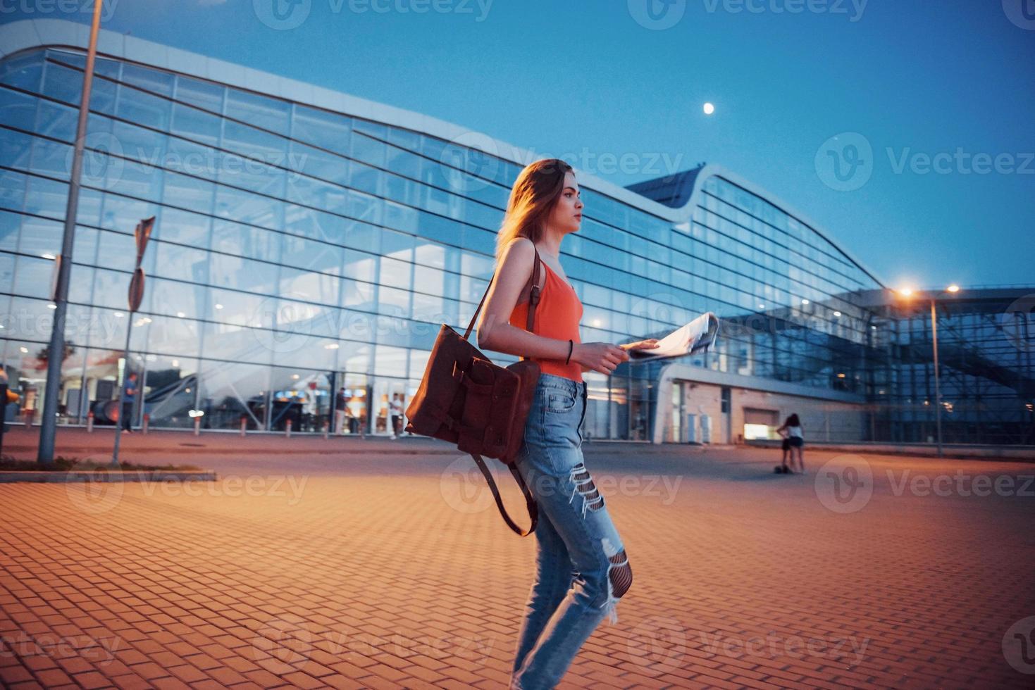 jeune fille coûte la nuit près du terminal de l'aéroport ou de la gare et lit le plan de la ville et cherche un hôtel. un touriste mignon avec des sacs à dos détermine le concept de voyage photo