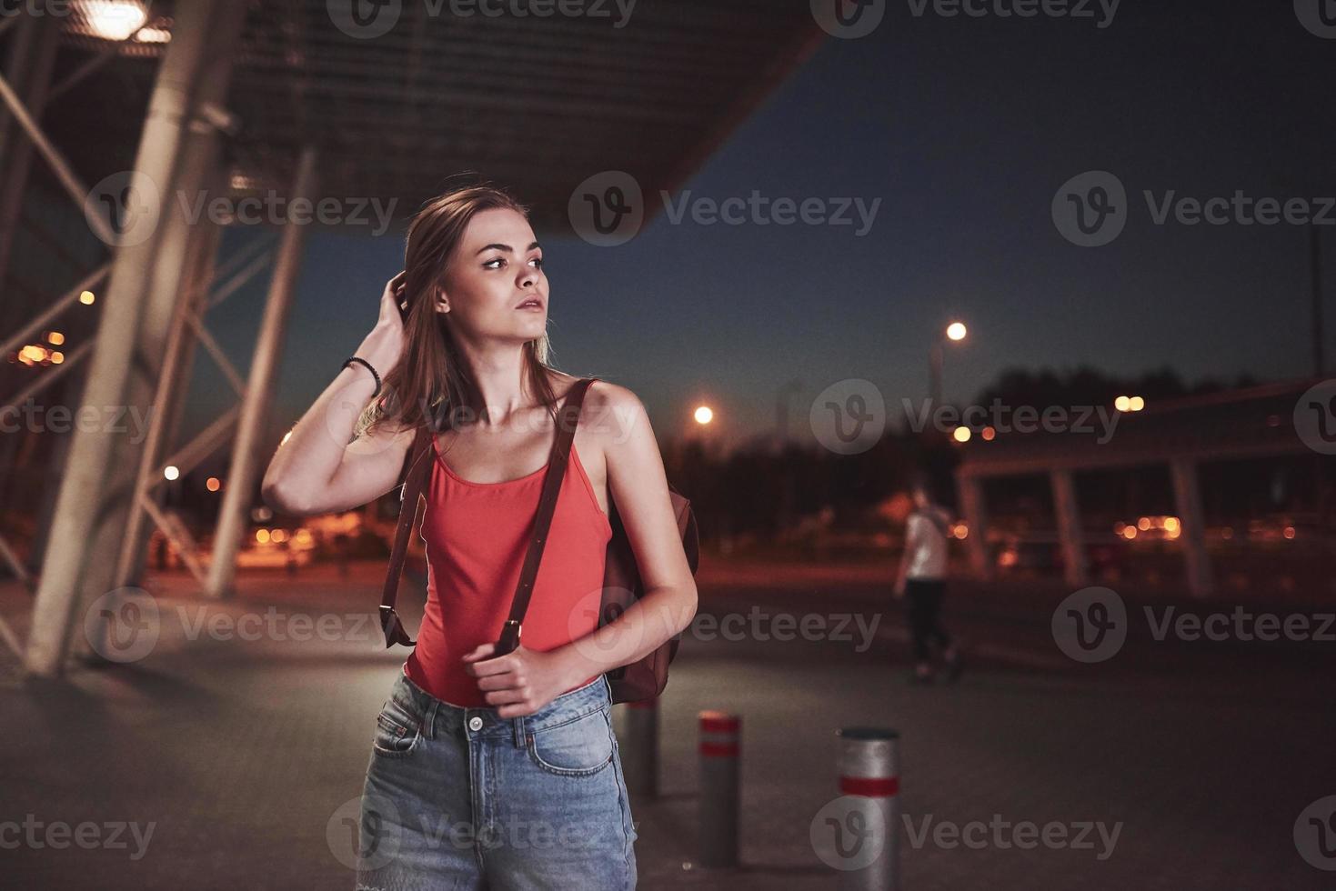 une belle jeune fille avec un sac à dos derrière ses épaules se tient dans la rue près d'un aéroport ou d'une gare par une chaude soirée d'été. elle vient d'arriver et attend un taxi ou ses amis photo