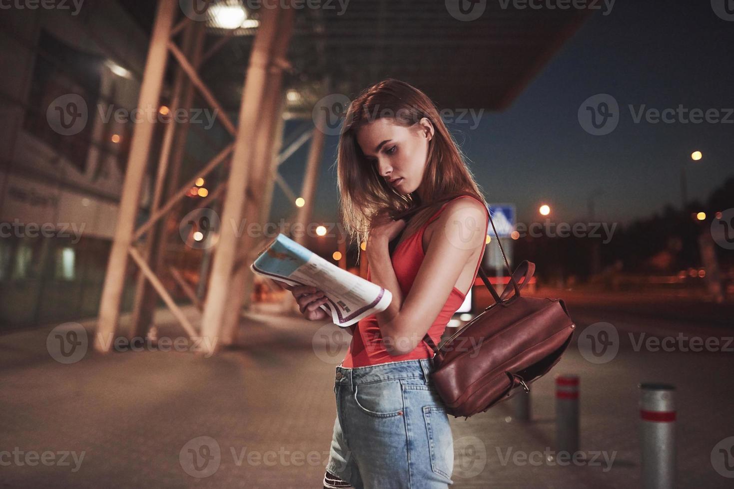 jeune fille coûte la nuit près du terminal de l'aéroport ou de la gare et lit le plan de la ville et cherche un hôtel. un touriste mignon avec des sacs à dos détermine le concept de voyage photo