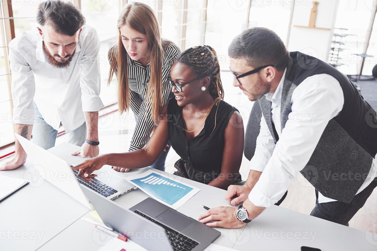 fille noire montrant des résultats et souriant. quatre employés multiraciaux travaillent au bureau en utilisant l'ordinateur portable sur la table photo