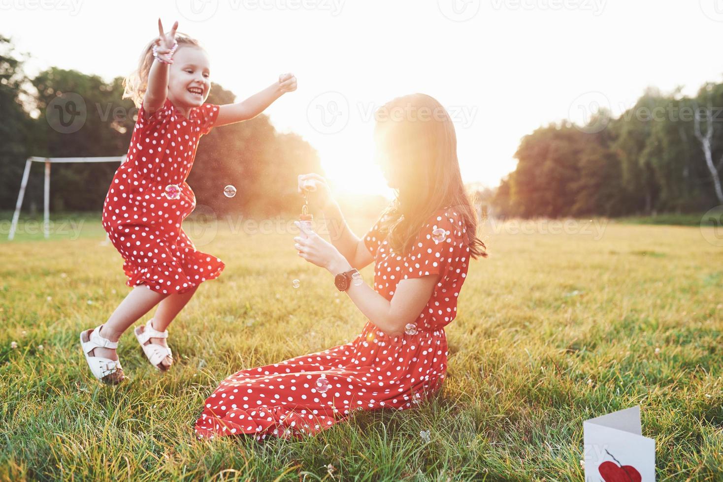 sauter avec les mains levées. fille avec sa fille s'amusant avec les bulles à l'extérieur assis sur l'herbe photo