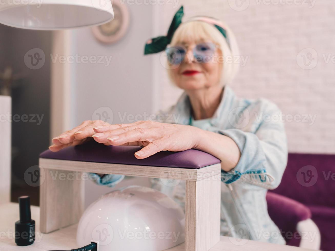 maître de manucure en gants bleus crémant les mains d'une femme élégante âgée en bleu lunettes de soleil et veste en jean assis au salon de manucure photo