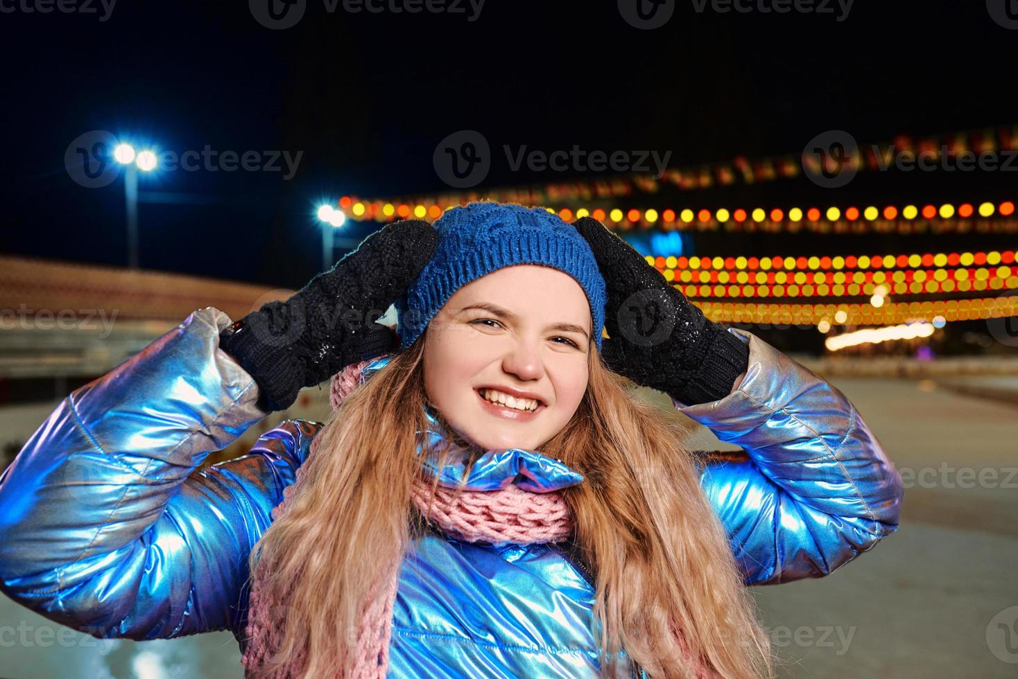 heureuse jeune femme caucasienne souriante en écharpe, chapeau, veste, mitaines près de la patinoire extérieure. nouvel an, amusement, concept d'hiver photo