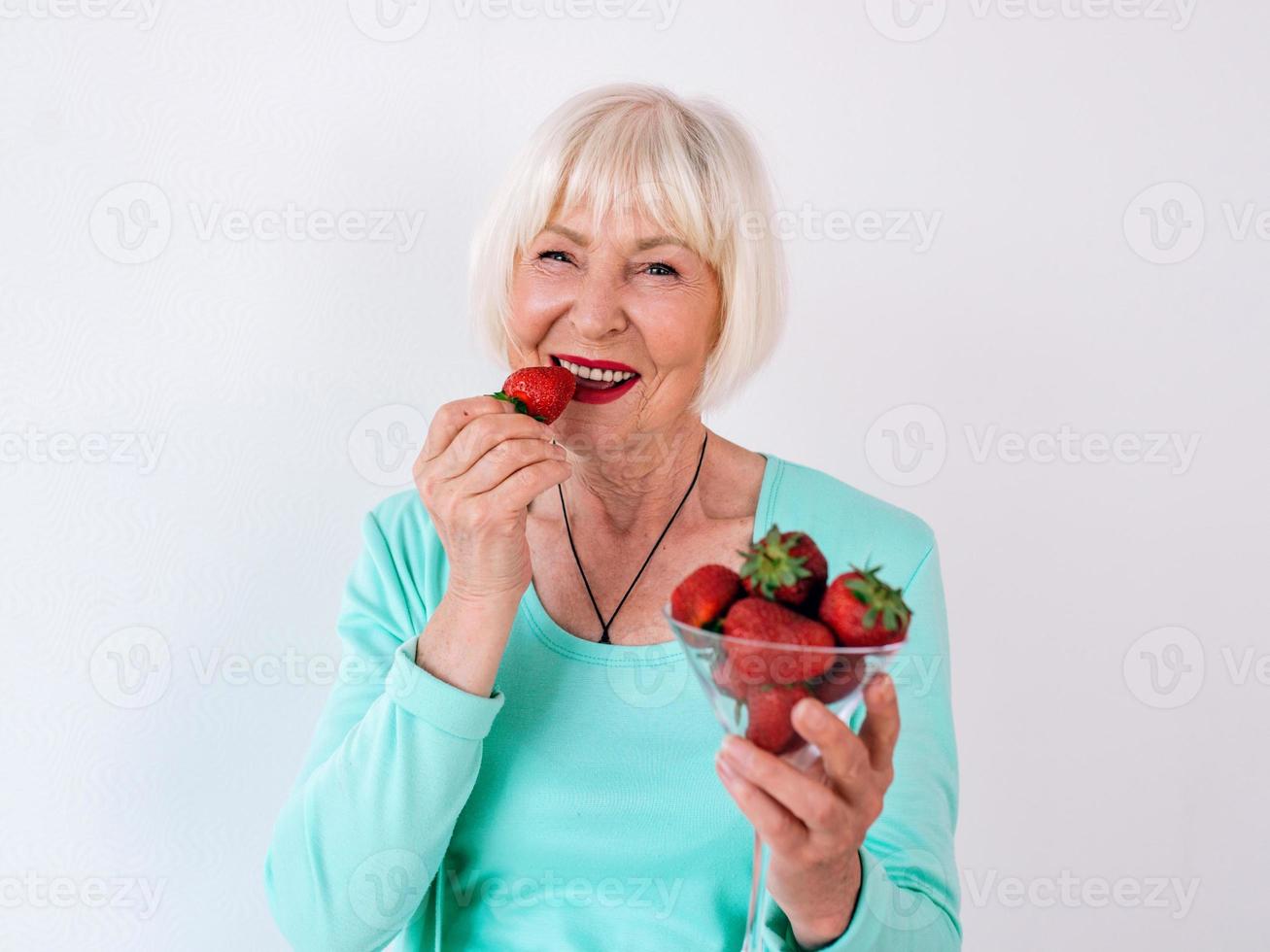 portrait d'une femme gaie élégante et âgée en vêtements turquoise mangeant des fraises. été, voyage, anti-âge, joie, retraite, fraises, baies, vitamines, concept de liberté photo