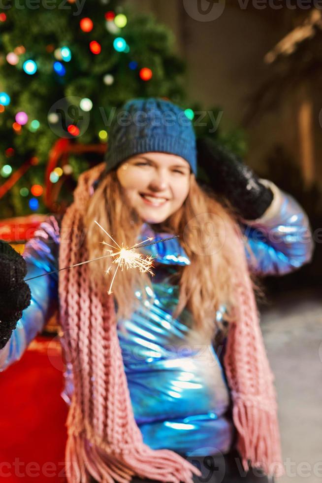 heureuse jeune femme caucasienne souriante en écharpe, chapeau, veste, mitaines avec cierge magique près de l'arbre de noël en plein air. nouvel an, amusement, concept d'hiver photo