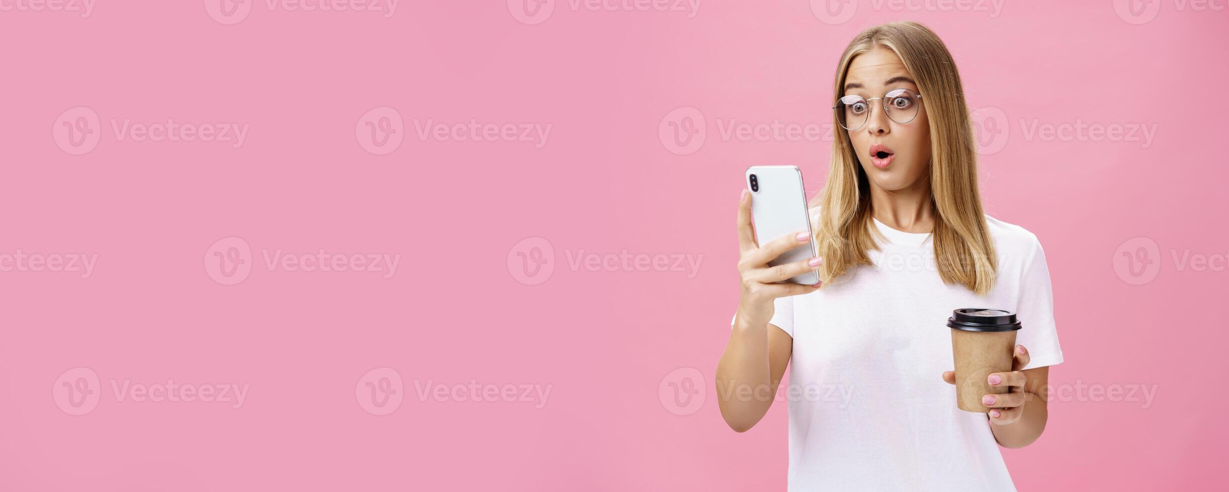 femme en buvant café étant sous le choc par reçu message réagir sur étourdissant nouvelles pliant lèvres halètement à la recherche étonné et impressionné à téléphone intelligent écran en portant papier tasse, posant plus de rose mur photo