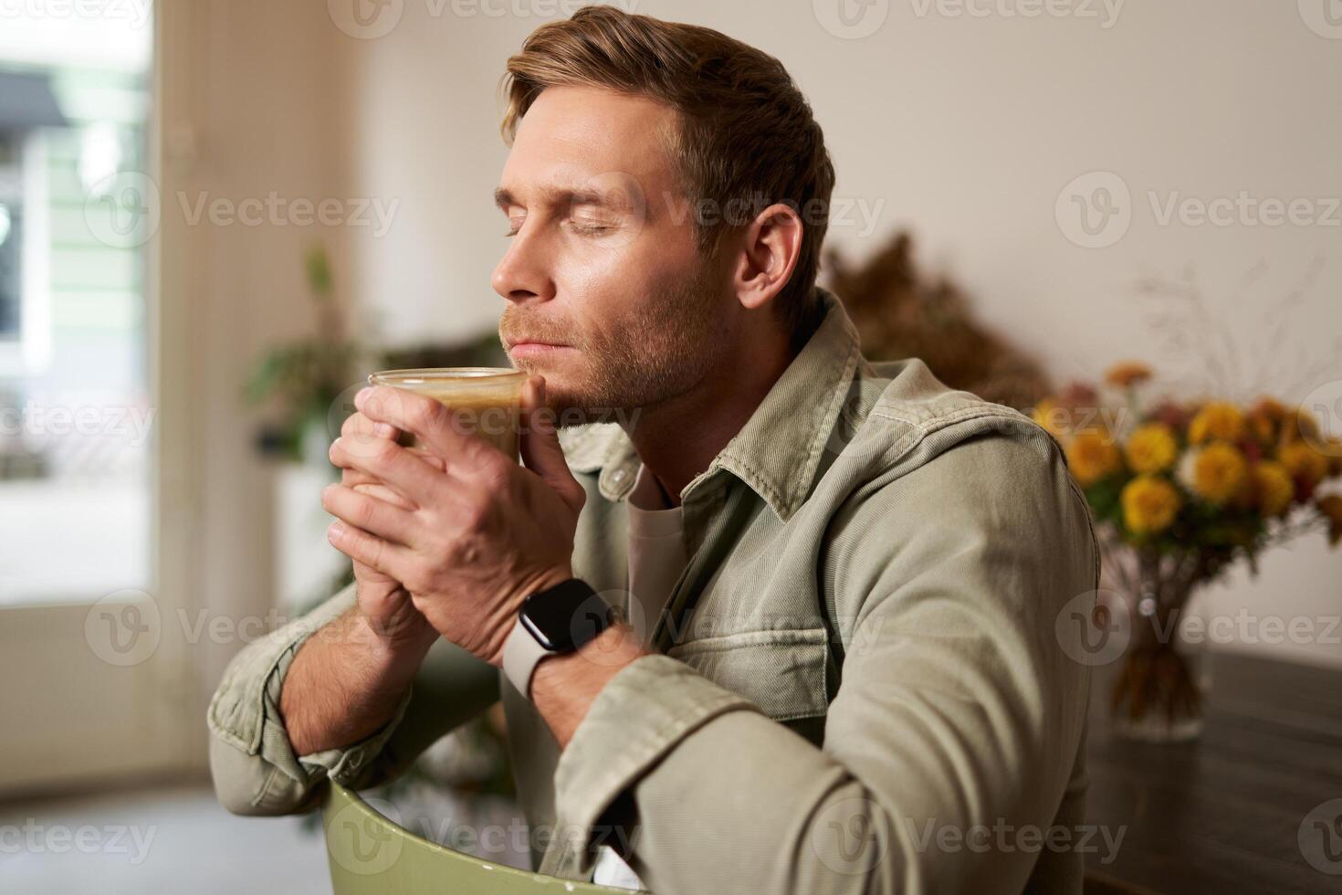 portrait de Jeune homme se ferme le sien yeux, les odeurs fraîchement fabriqué café dans café, est assis sur chaise avec verre de cappuccino, jouit le goût photo