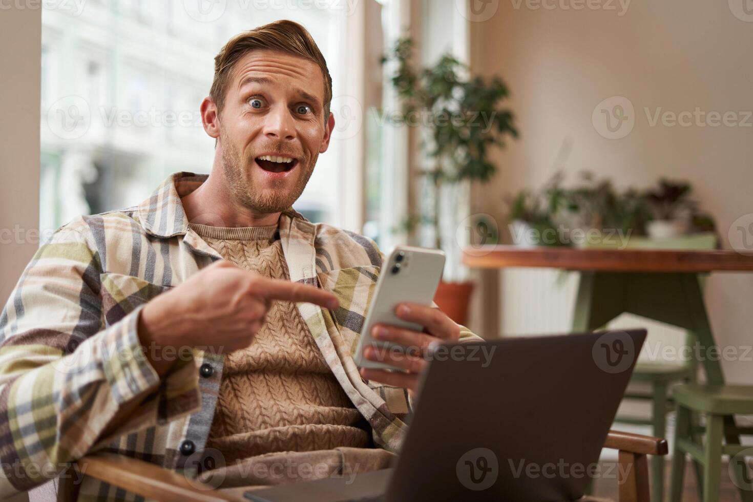 excité gars dans café, séance avec téléphone et montrer du doigt à écran avec intérêt, recommander un application, en utilisant mobile dispositif et portable dans cotravail espace. concept de free-lance et éloigné espace de travail photo