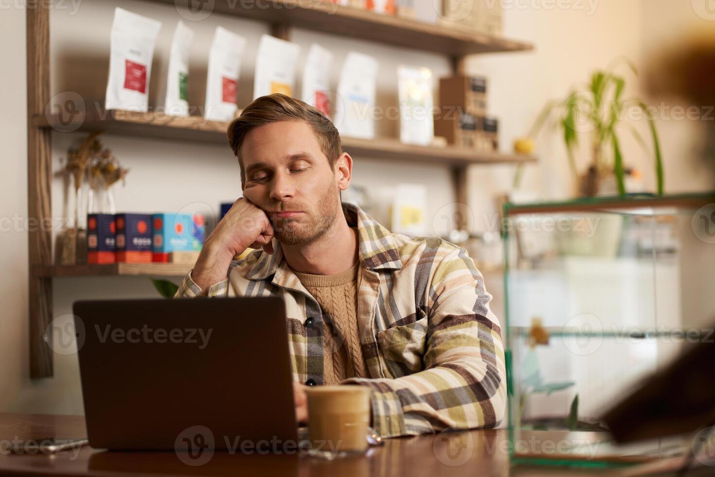 portrait de ennuyé pigiste, numérique nomade séance avec portable dans café, penché sur le sien poing, à la recherche avec ennui à ordinateur filtrer, en buvant café, Faire travail tâche photo