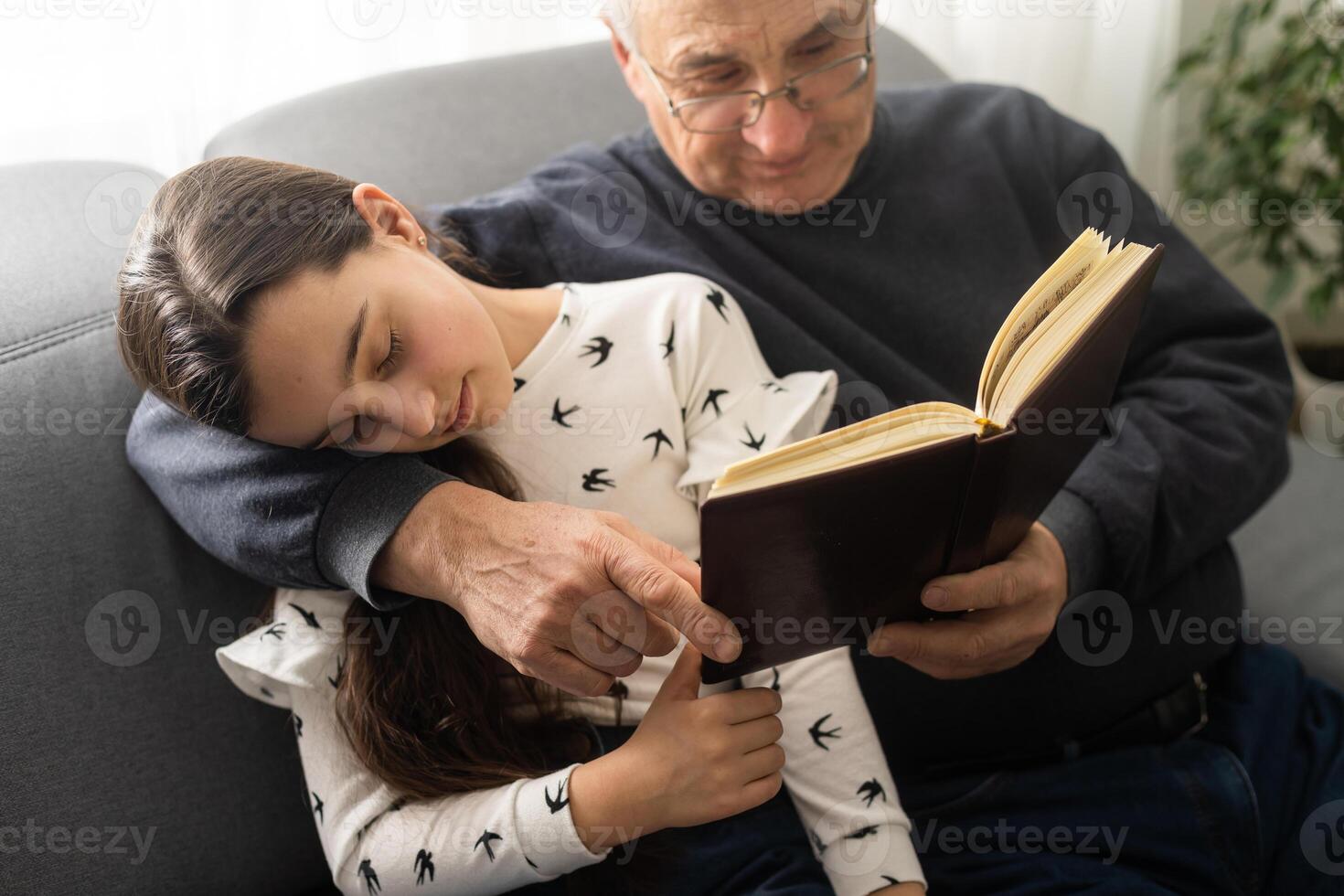 photo de vieilli vieux grand-père peu jolie petite fille asseoir canapé étreindre rester maison quarantaine sécurité lis intéressant livre Fée conte moderne intérieur vivant pièce à l'intérieur.