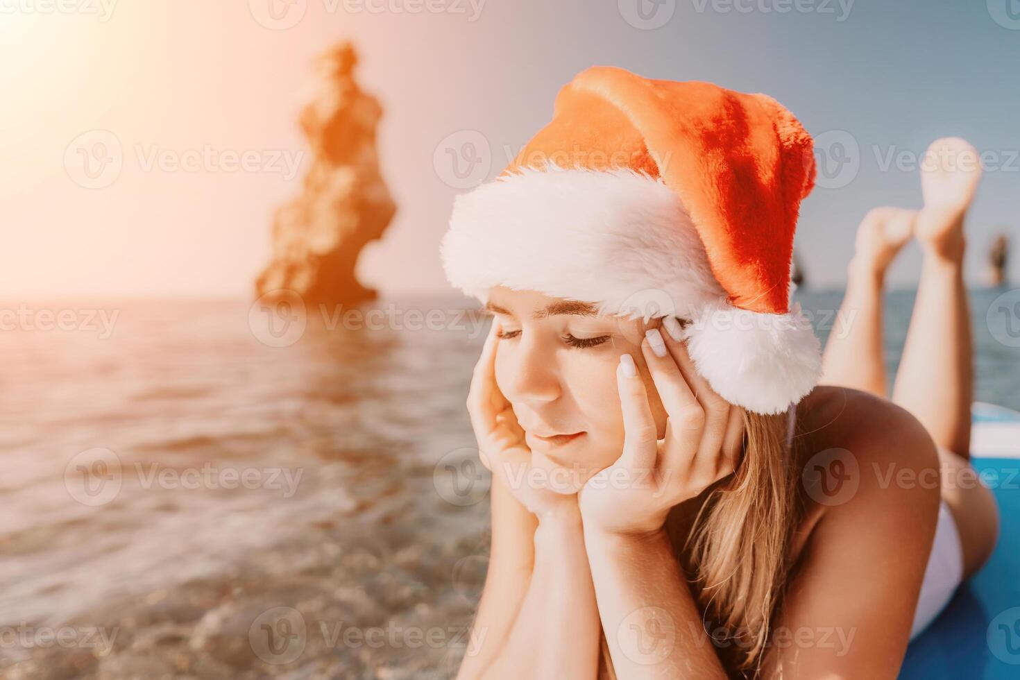 femme mer souper. proche en haut portrait de content Jeune caucasien femme avec longue cheveux dans Père Noël chapeau à la recherche à caméra et souriant. mignonne femme portrait dans une blanc bikini posant sur souper planche dans le mer photo