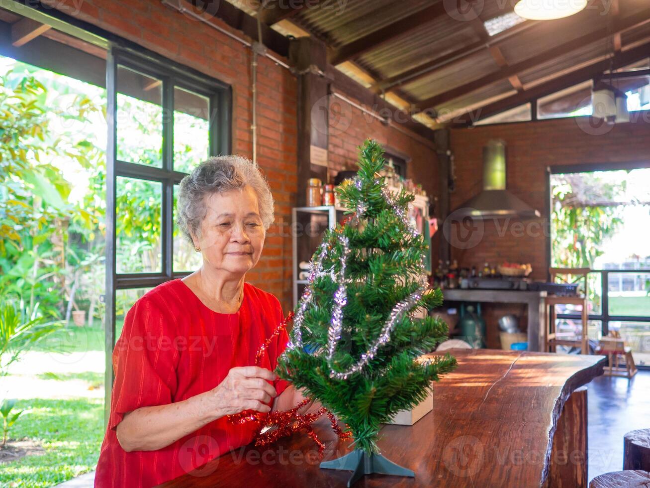 Sénior femme décorer le Noël arbre avec blanc et rouge rubans tandis que séance sur une chaise à maison. concept de vieilli gens et Noël et Nouveau année Festival photo