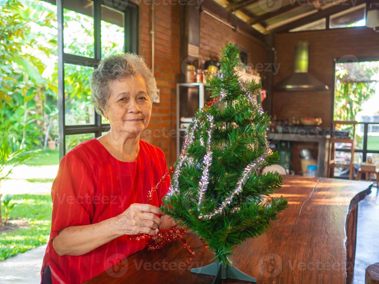 Sénior asiatique femme décorer le Noël arbre avec blanc et rouge rubans à maison. concept de vieilli gens et Noël et content Nouveau année Festival photo