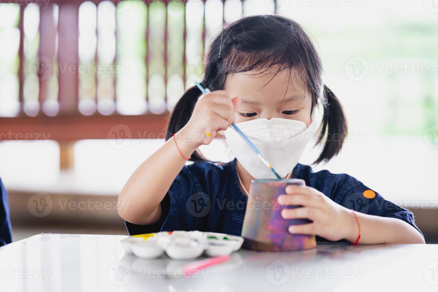 enfant faisant de l'artisanat avec un pinceau. petite fille portant un masque chirurgical blanc pour empêcher la propagation du virus et de la pollution par la poussière pm2.5. photo