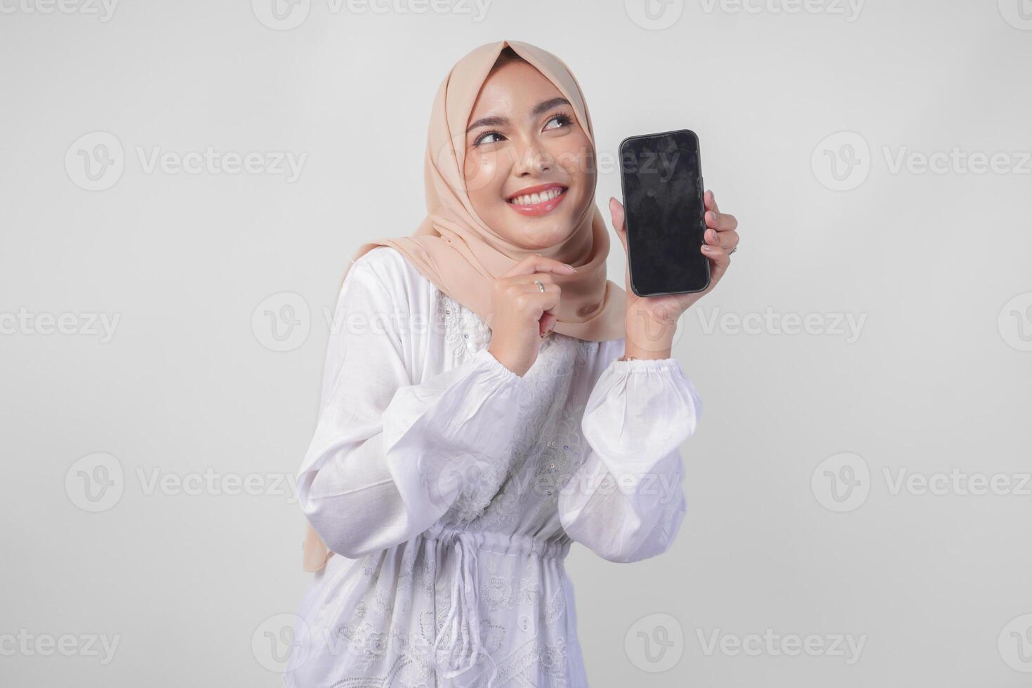 portrait de Jeune asiatique musulman femme portant blanc robe et hijab souriant Heureusement, montrer du doigt à le Vide écran copie espace sur sa téléphone et montrant écran à le caméra photo