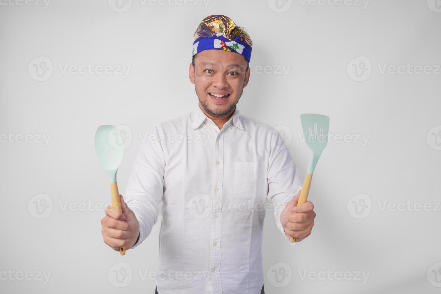 souriant Jeune balinais homme dans blanc chemise et traditionnel coiffure en portant spatule et cuisine cuisine ustensiles photo