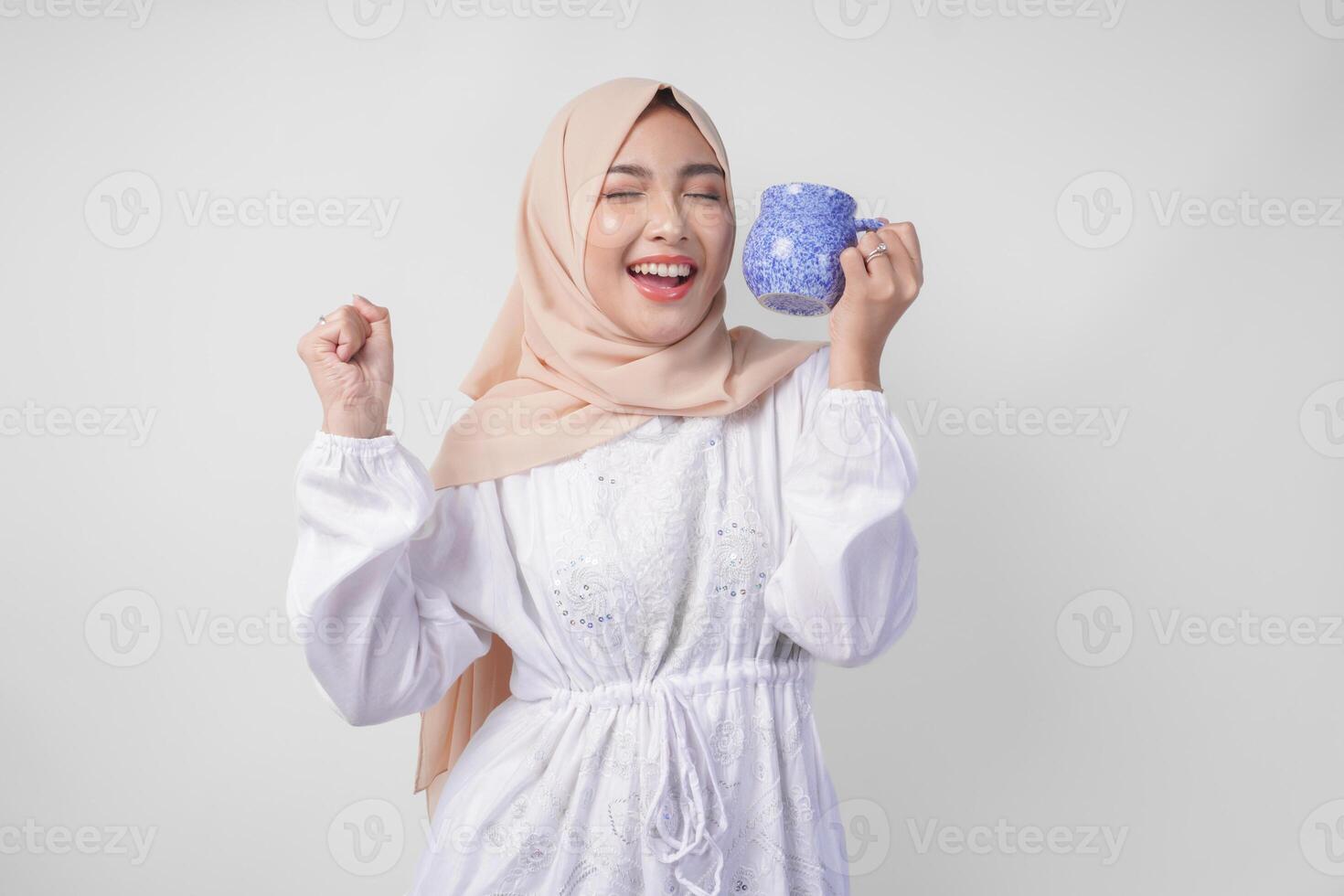 joyeux asiatique musulman femme portant blanc robe et hijab en portant une verre de l'eau dans une bleu tasse avec une content réussi geste, sentiment soulagé après jeûne, isolé par blanc Contexte photo