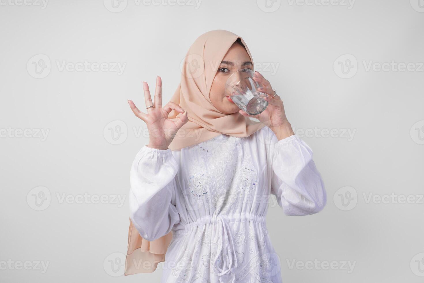 Jeune asiatique musulman femme dans blanc robe et hijab en portant et en buvant tasse de Frais l'eau tandis que fabrication un d'accord, bien, agréable signe geste en utilisant les doigts photo