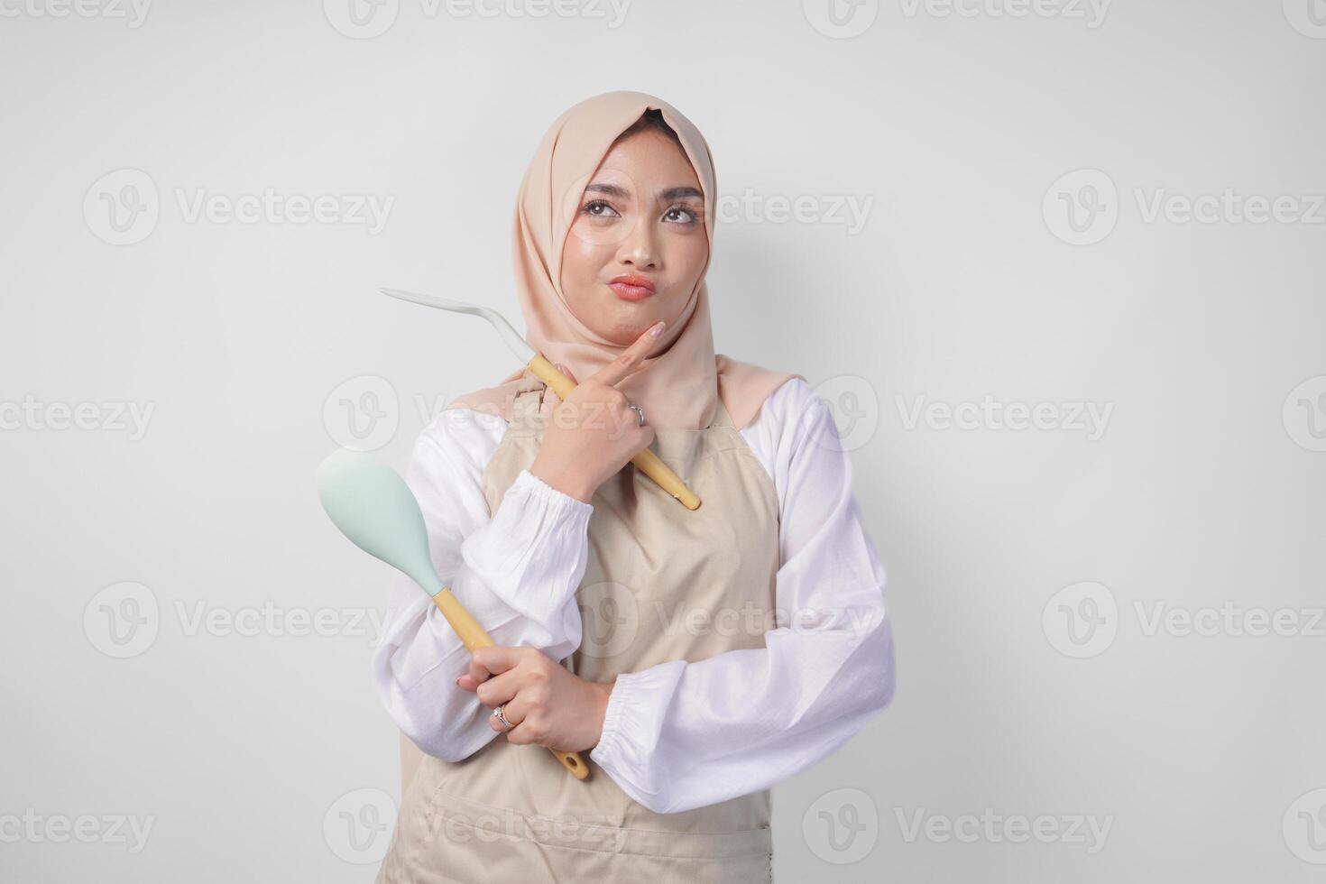 réfléchi Jeune asiatique musulman femme dans hijab et crème tablier en portant spatule et cuisine cuisine ustensiles, en pensant difficile quoi nourriture menu à cuisinier photo
