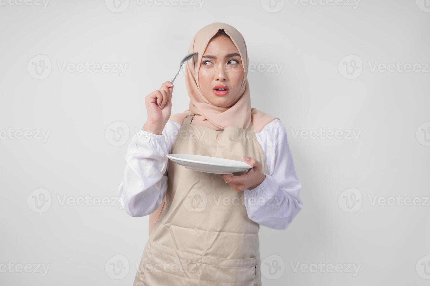 confus Jeune asiatique musulman femme dans hijab et crème tablier en portant cuillère et vide assiette avec copie espace plus de il, en pensant difficile quoi nourriture menu à manger pour iftar. Ramadan concept photo