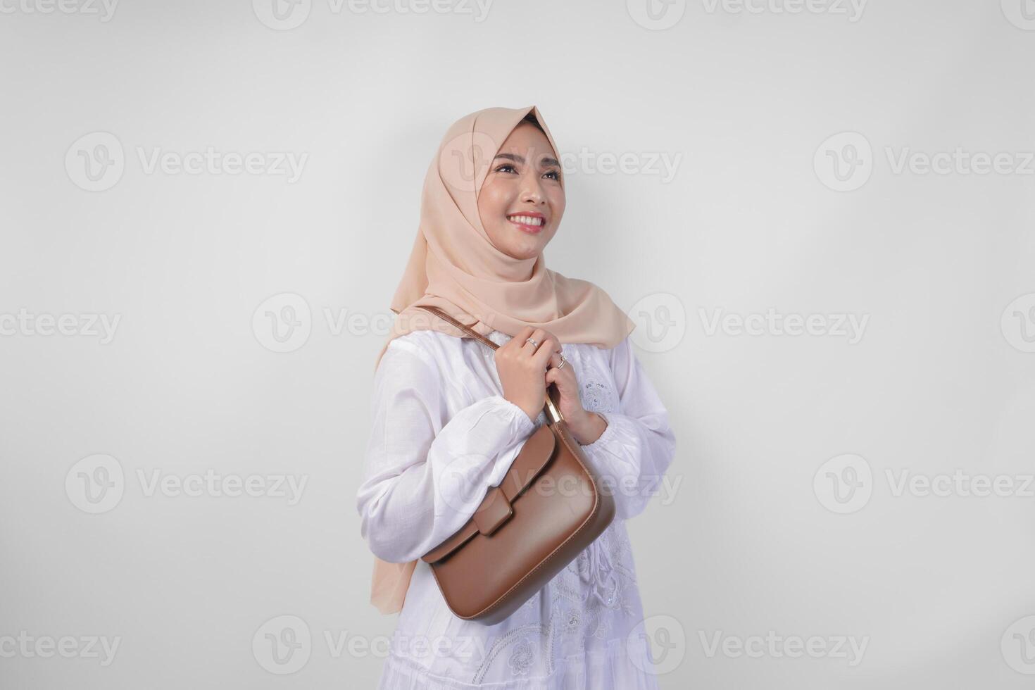 élégant Jeune asiatique musulman femme portant blanc robe et hijab avec marron fronde sac souriant Heureusement plus de isolé blanc Contexte. ramadhan mode concept photo