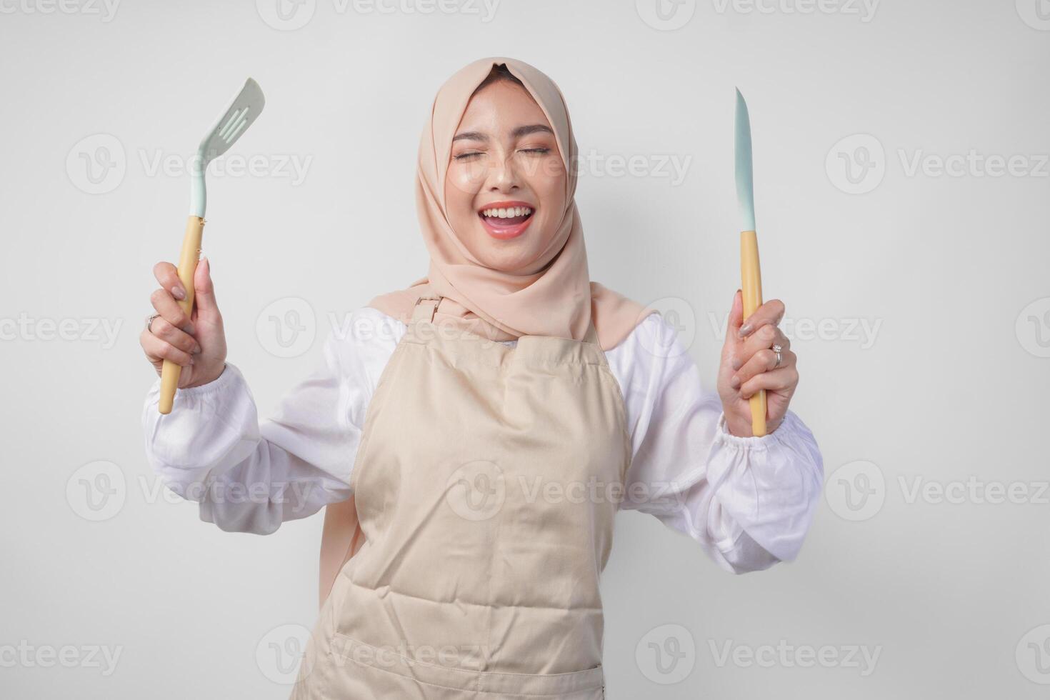 excité Jeune asiatique musulman femme dans une voile hijab et crème tablier souriant à le caméra tandis que en portant spatule et cuisine cuisine ustensiles photo