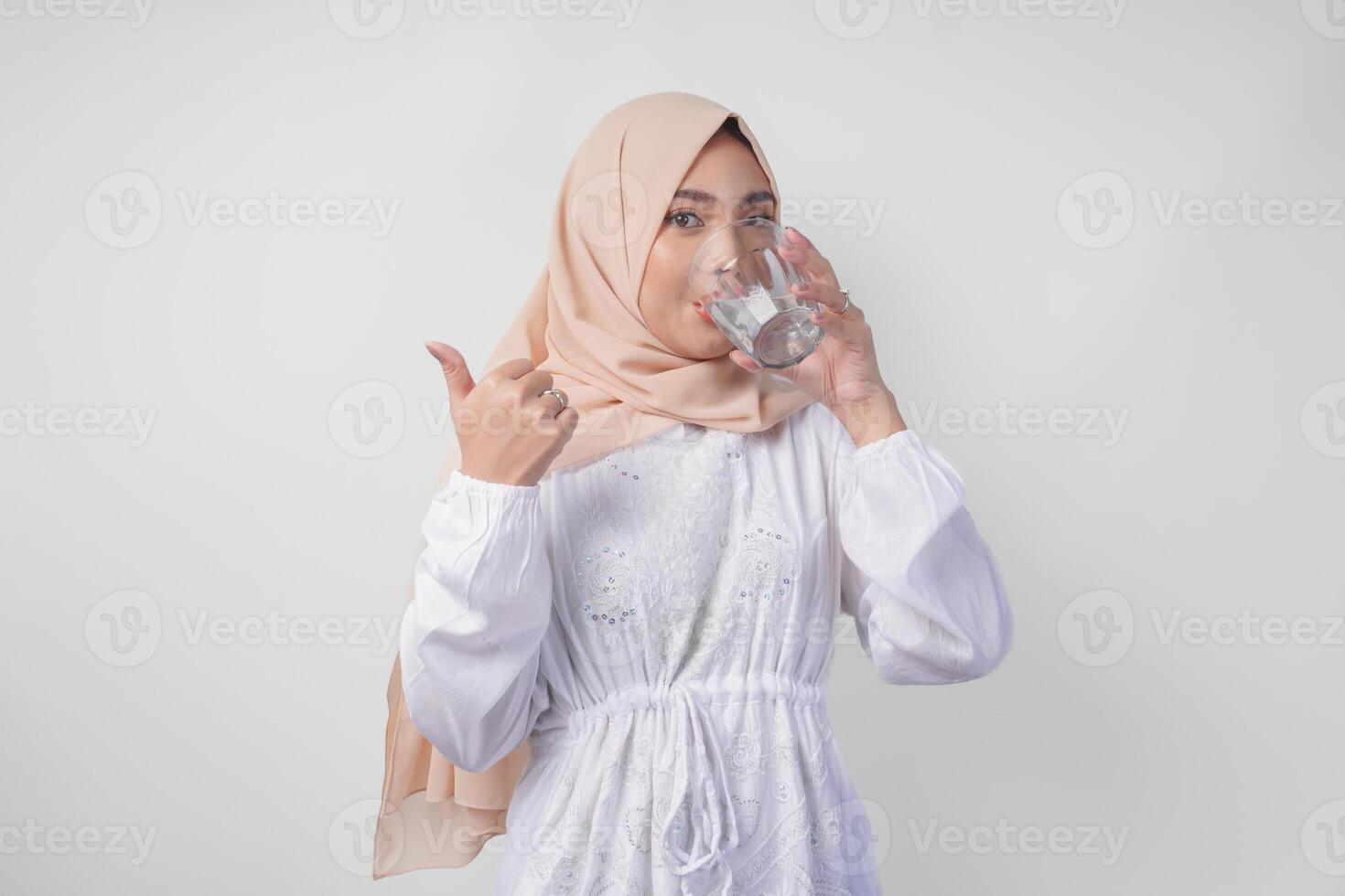 soif Jeune asiatique musulman femme dans blanc robe et crème voile hijab en buvant Frais minéral l'eau après jeûne tandis que Faire les pouces en haut geste. Ramadan concept photo