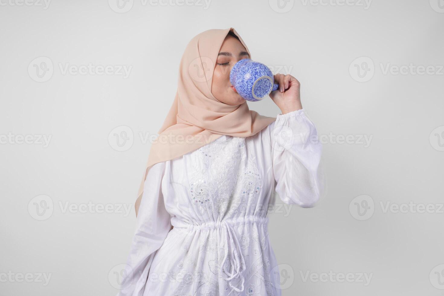 soif Jeune asiatique musulman femme dans blanc robe et crème voile hijab en buvant Frais minéral l'eau de une bleu tasse après jeûne. Ramadan concept photo