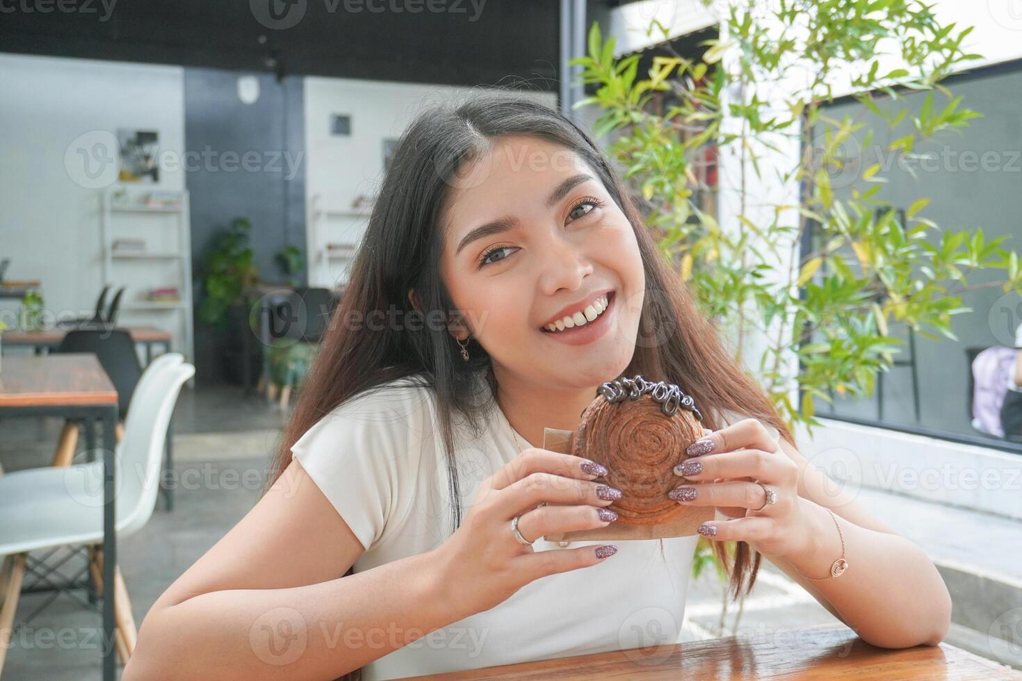 Jeune asiatique femme en portant et montrant une noir assiette de Donut Pâtisserie nommé cromboloni avec Chocolat remplissage pour casse-croûte temps, souriant avec une content et de bonne humeur expression photo