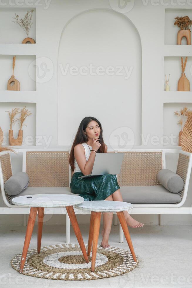 réfléchi Jeune femme en utilisant portable tandis que séance sur une canapé concentré et dans Profond pensée, travail sur vacances photo