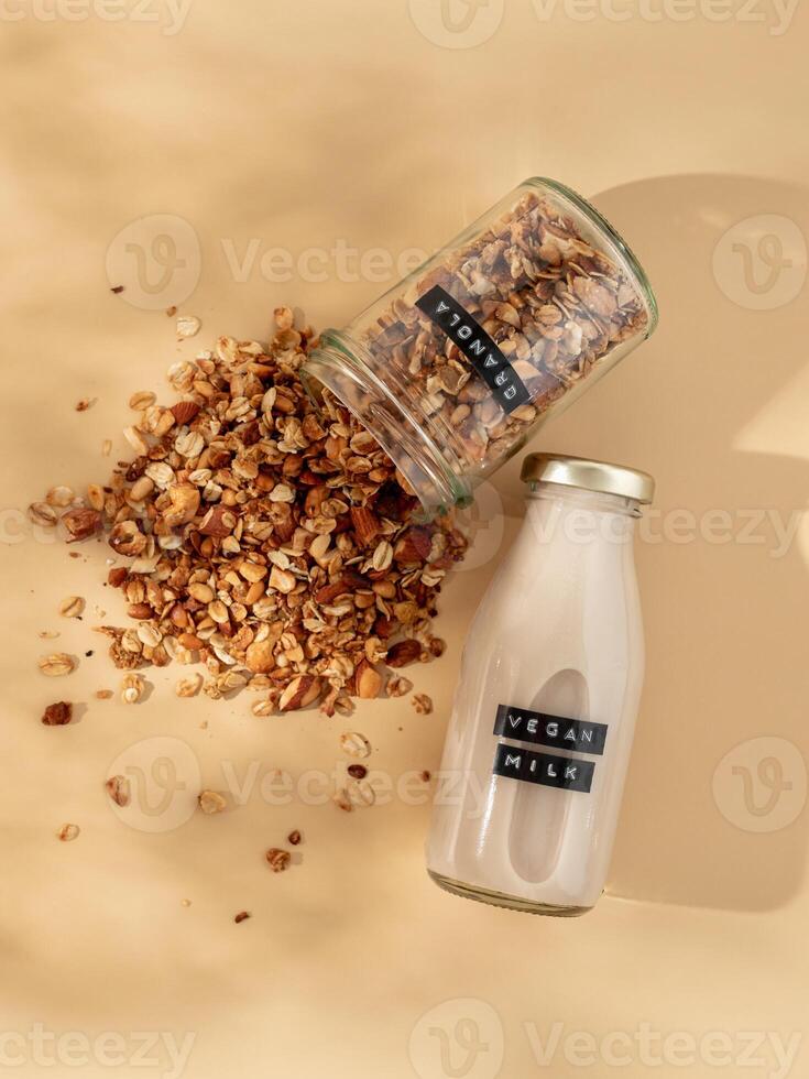 granola et végétalien lait. en bonne santé petit déjeuner esthétique concept. fait maison granola saupoudré de verre pot et végétalien Lait dans verre bouteille plus de Champagne Jaune ou pastel beige Contexte. Haut vue plat allonger photo