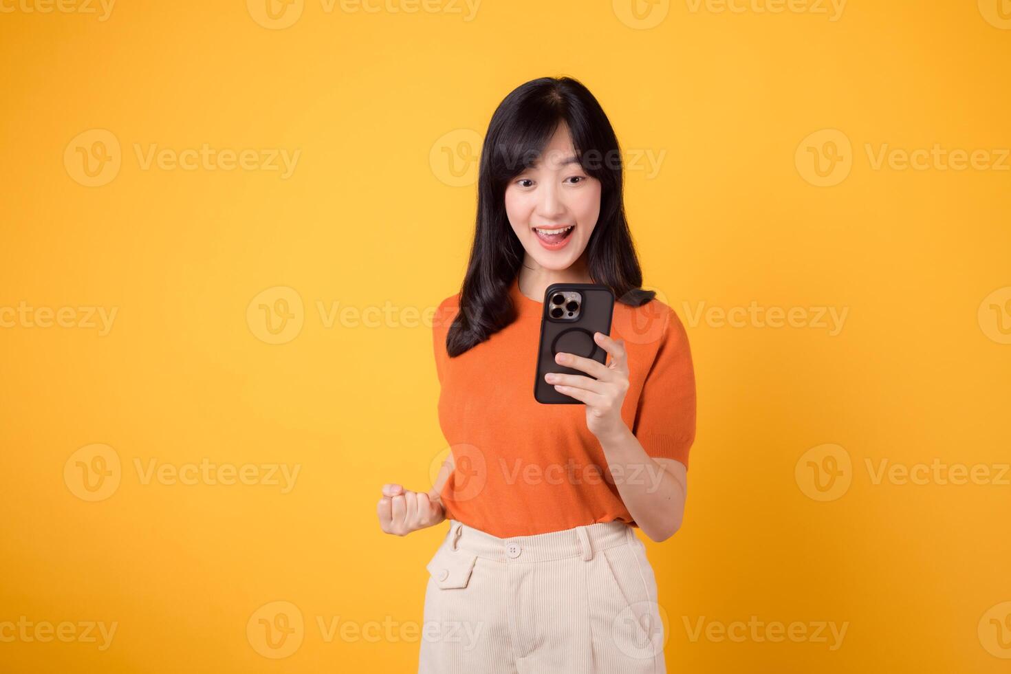 souriant asiatique femme dans 30s, portant Orange chemise, en utilisant téléphone intelligent avec poing en haut main signe sur vibrant Jaune Contexte. célébrer Nouveau app concept photo