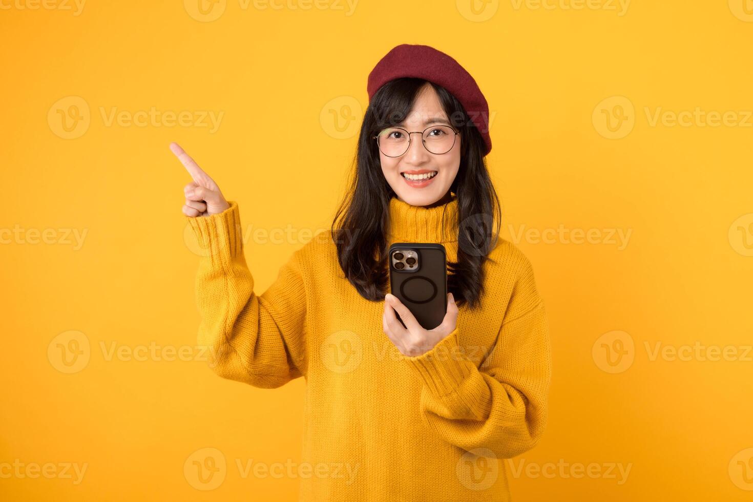 portrait Jeune magnifique 30s asiatique femme content sourire portant Jaune chemise et rouge chapeau montrer du doigt doigt geste à gratuit copie espace et en utilisant téléphone intelligent contre Jaune Contexte. Nouveau application photo