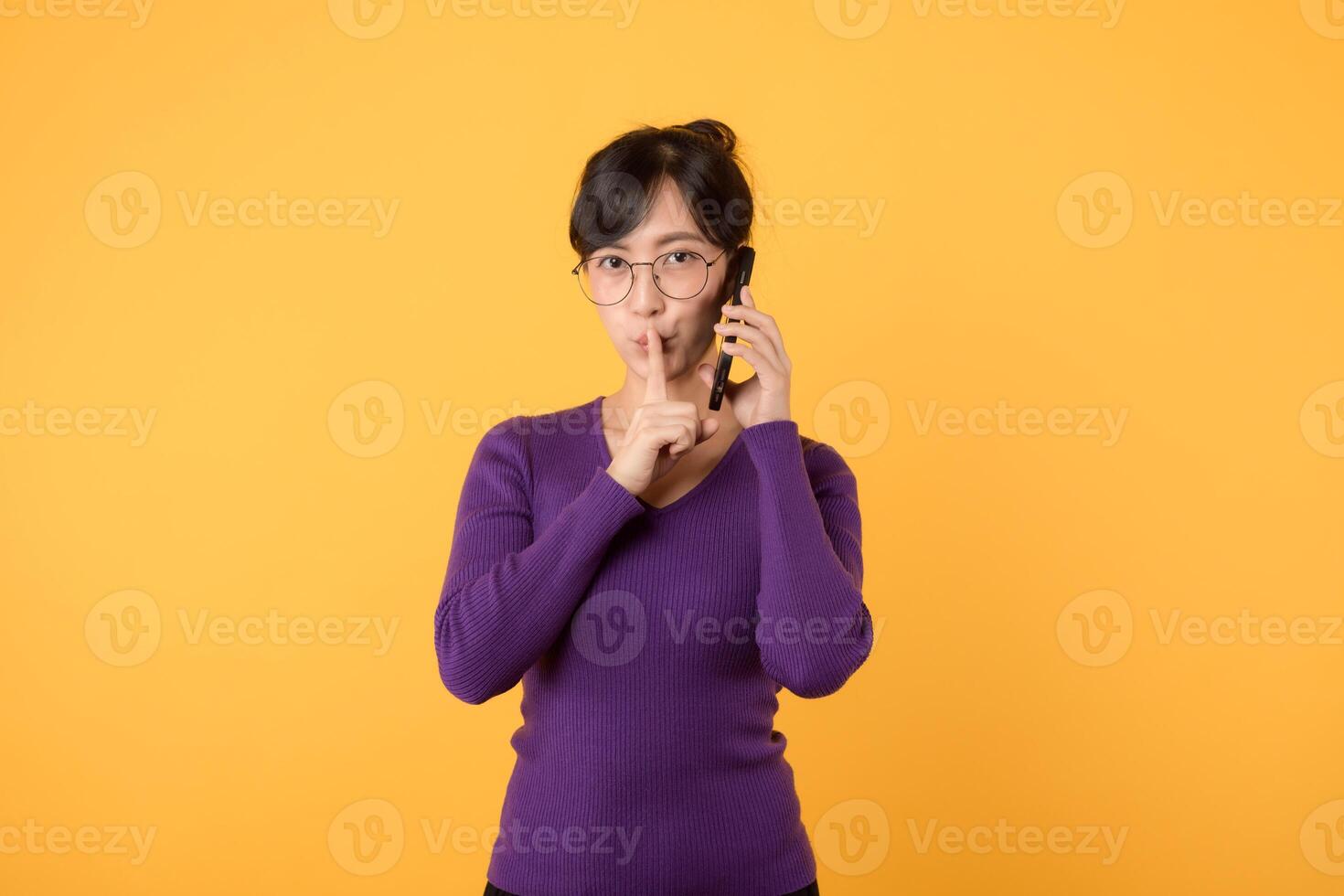 mystérieux asiatique femme avec violet chemise fait du silence geste presses indice doigt à lèvres actions secret avec vous détient mobile téléphone porte optique lunettes et chaud sauteur isolé plus de Jaune mur photo