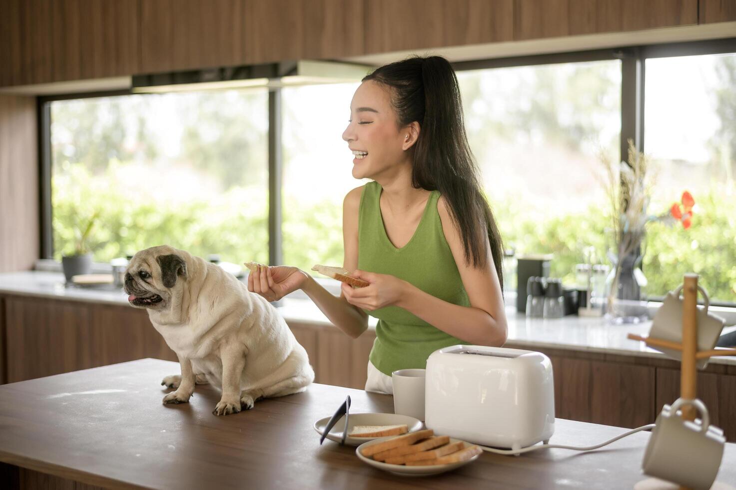 asiatique femme en train de préparer café et pain grillé pain pour petit déjeuner prendre plaisir avec chien à le cuisine table dans le Matin photo