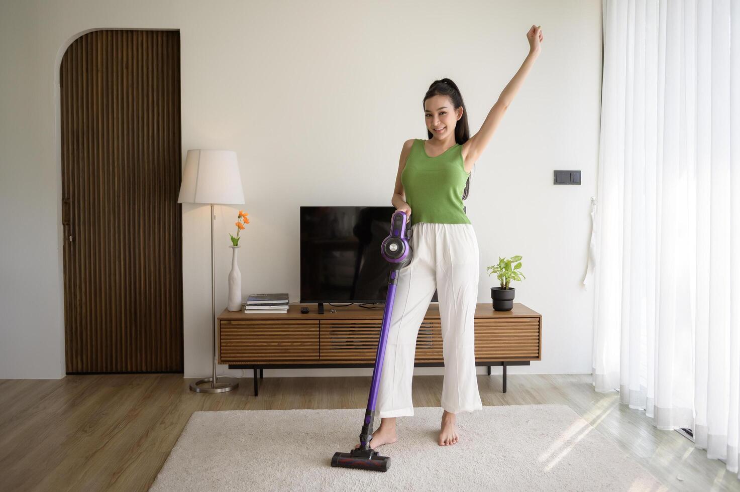 Jeune content femme en utilisant une vide nettoyeur le sol dans vivant pièce photo