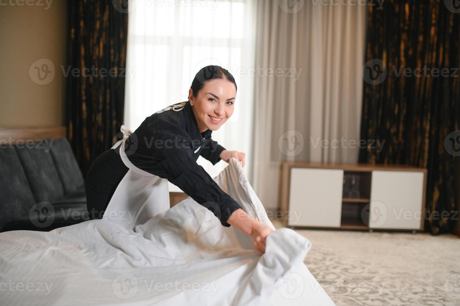 Jeune jolie gouvernante dans uniforme en changeant draps dans Hôtel pièce tandis que permanent par double lit photo