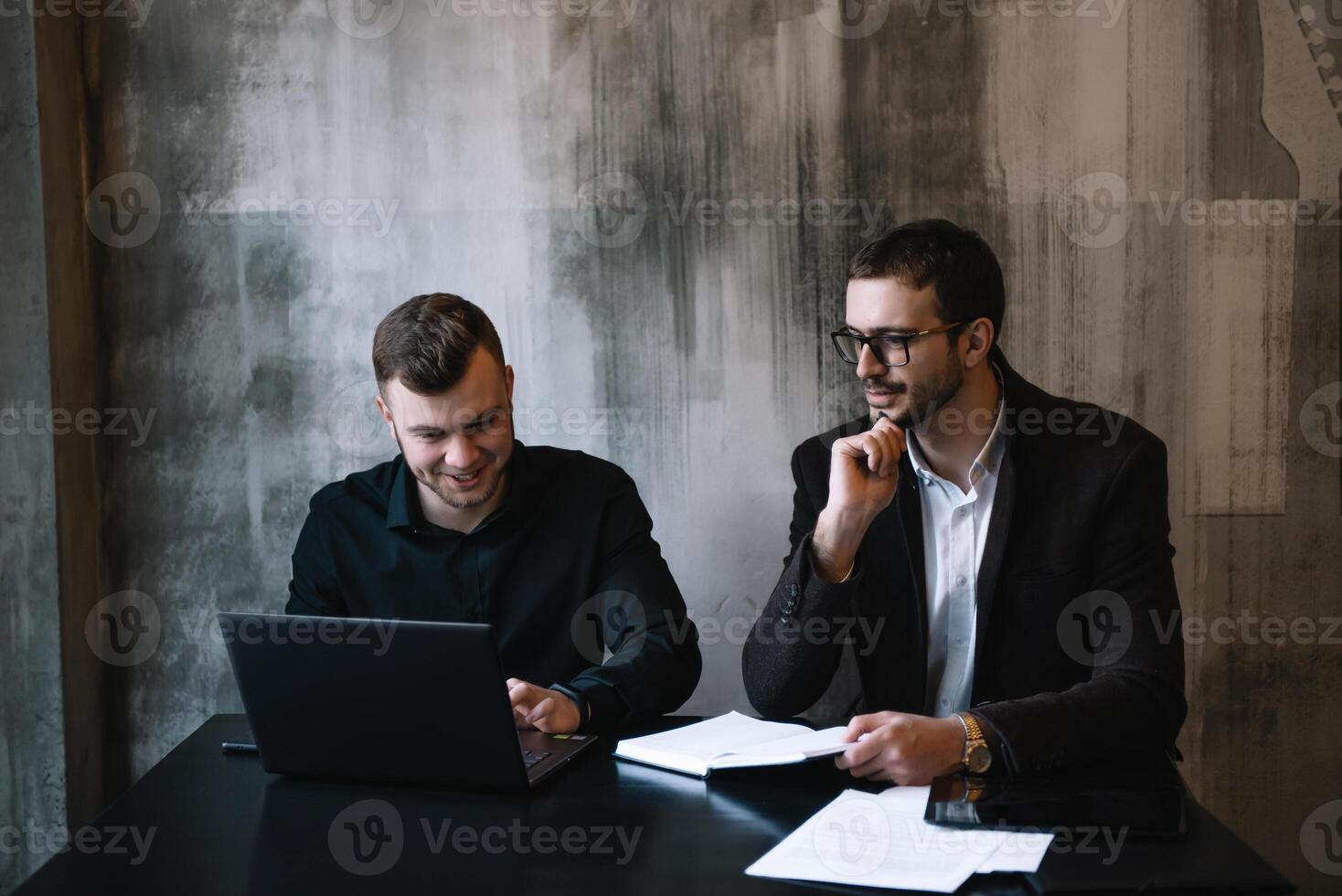 deux les hommes d'affaires dans un Bureau souriant à le caméra tandis que travail ensemble derrière une portable ordinateur. photo