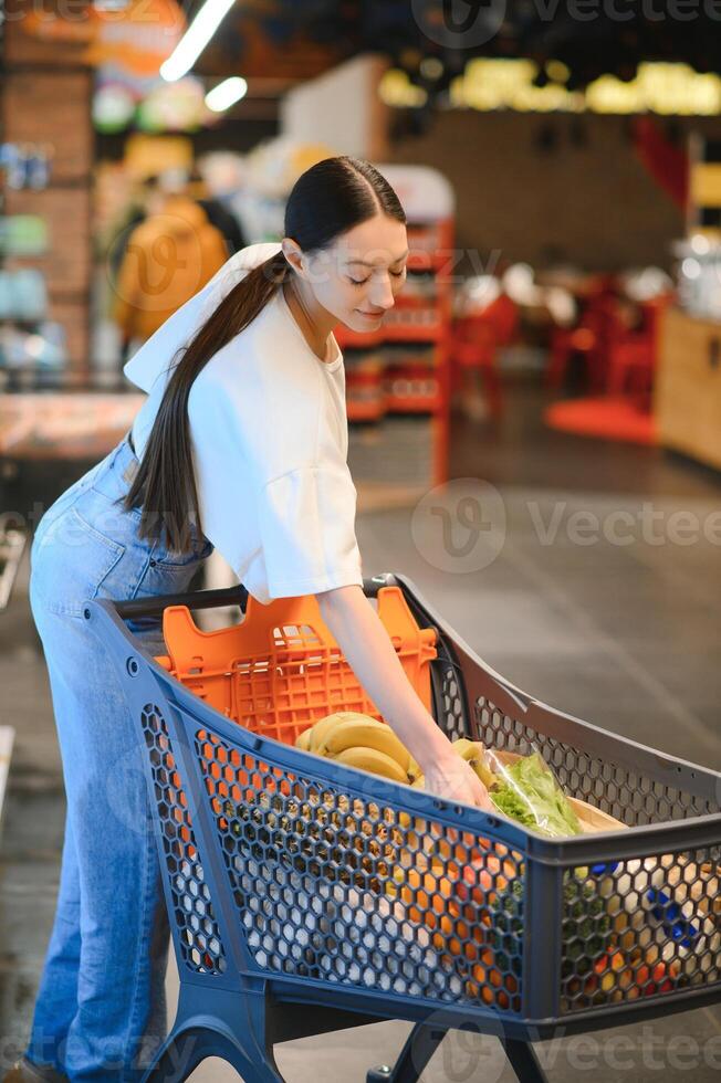 Jeune souriant content femme Années 20 dans décontractée vêtements achats à supermarché boutique avec épicerie Chariot photo