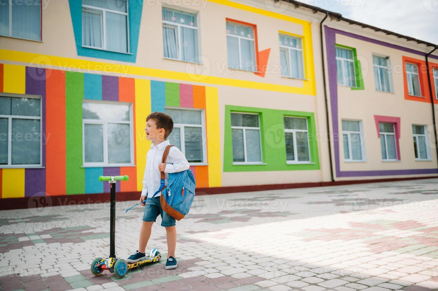 adolescent garçon avec donner un coup scooter près moderne école. enfant avec sac à dos et livre en plein air. début de cours. premier journée de automne. retour à école. photo