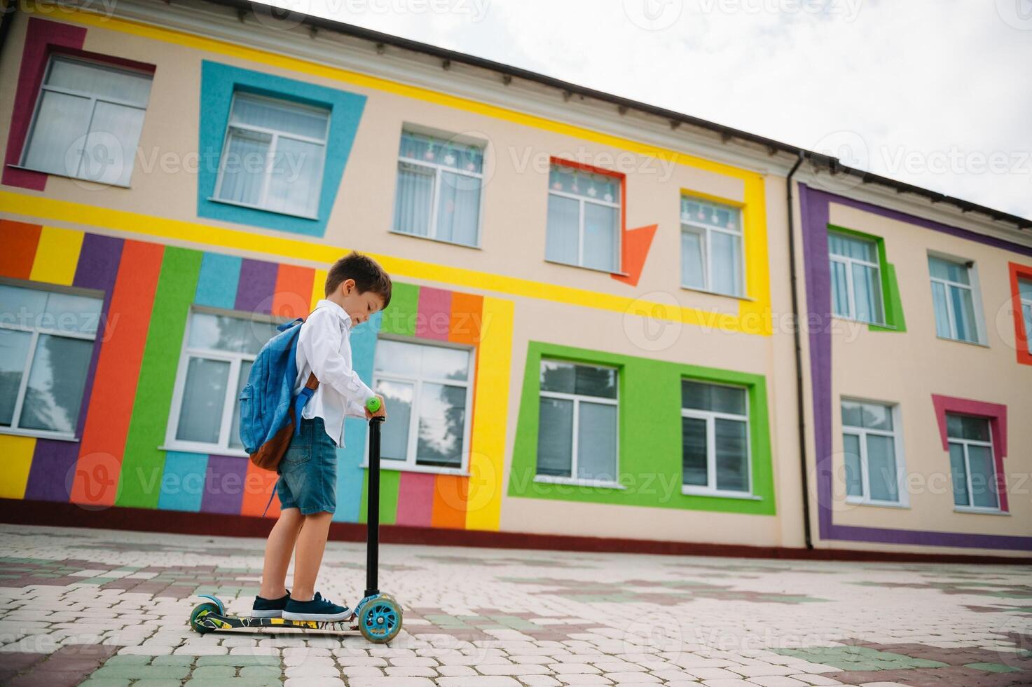 adolescent garçon avec donner un coup scooter près moderne école. enfant avec sac à dos et livre en plein air. début de cours. premier journée de automne. retour à école. photo