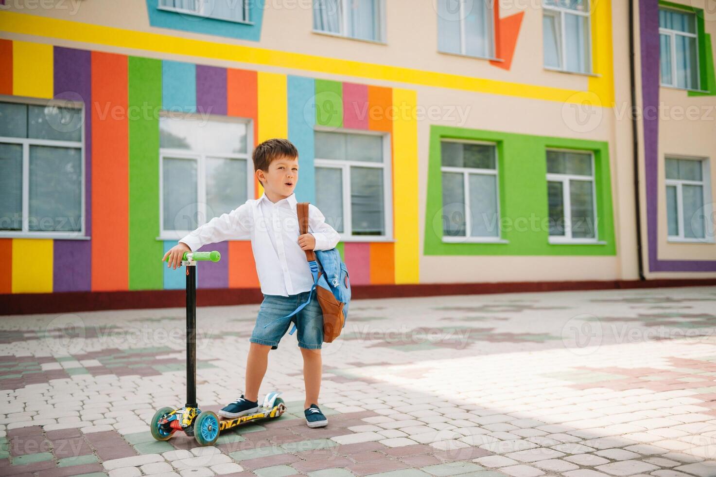 adolescent garçon avec donner un coup scooter près moderne école. enfant avec sac à dos et livre en plein air. début de cours. premier journée de automne. retour à école. photo