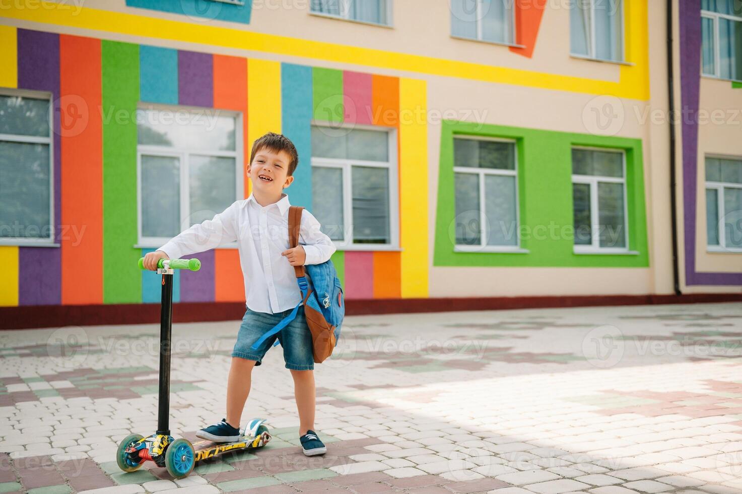 adolescent garçon avec donner un coup scooter près moderne école. enfant avec sac à dos et livre en plein air. début de cours. premier journée de automne. retour à école. photo